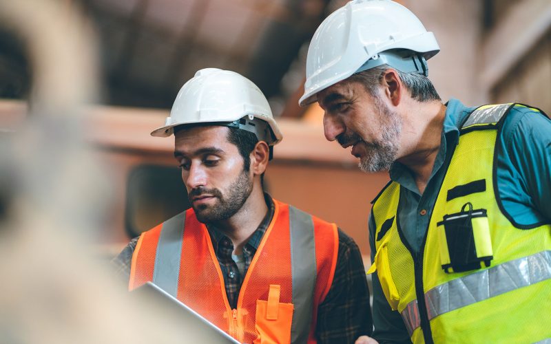 Teamwork men in construction site, Two civil engineer or professional foreman in safety helmet hard hat using blueprint in digital tablet working while standing at industrial factory, teamwork concept